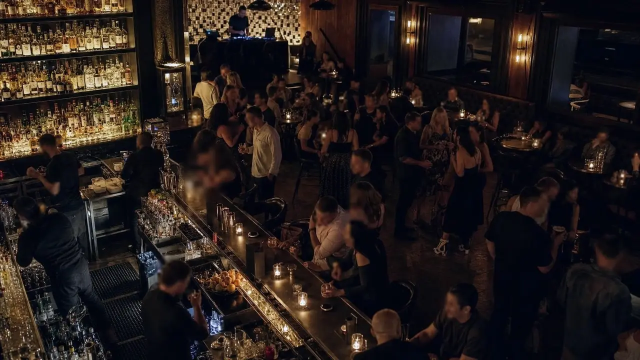 The interior of a bar viewed from above the bar, with a tall backlit spirits shelf, candlelit tables, and people standing and seated in groups around an open floor