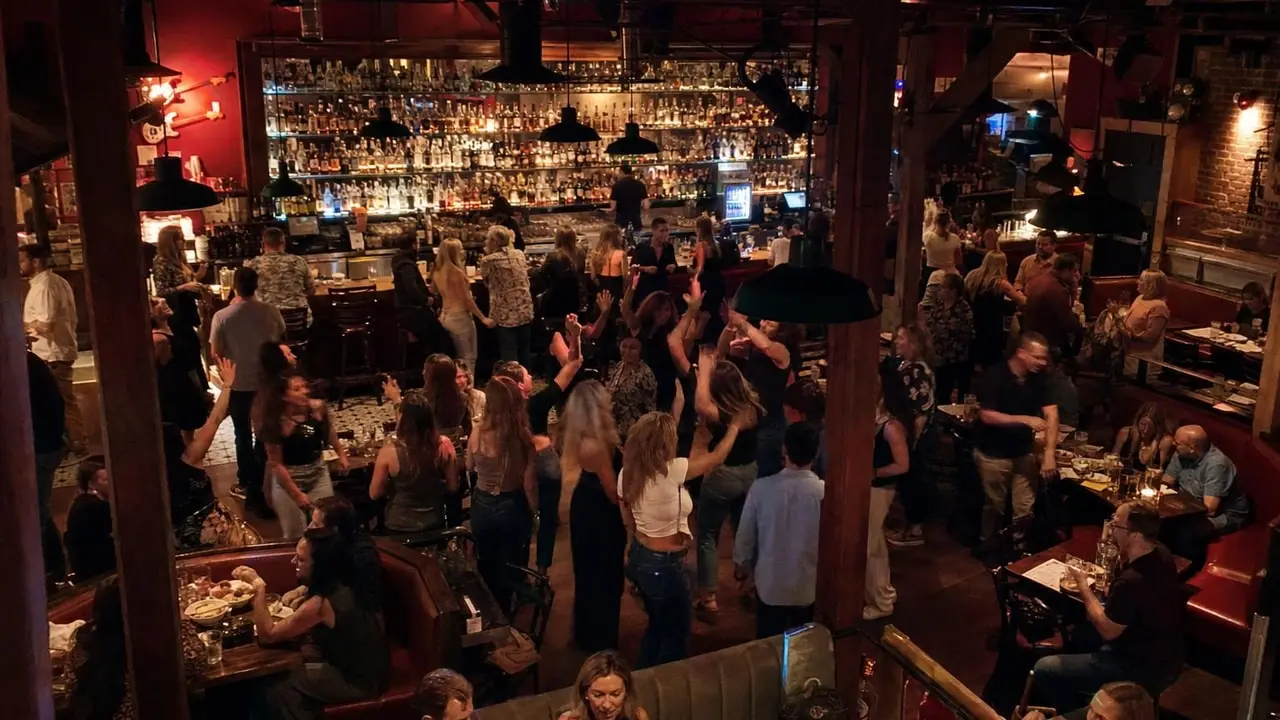 A crowded bar room at night, with people dancing under warm light in front of a glowing back bar