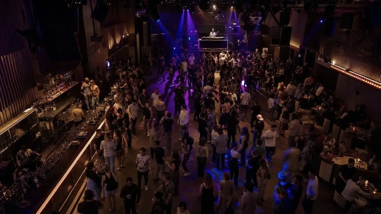 The interior of a music venue viewed from above, with coloured stage lighting across a dance floor, a long bar along the left side, and groups of people separated by open space